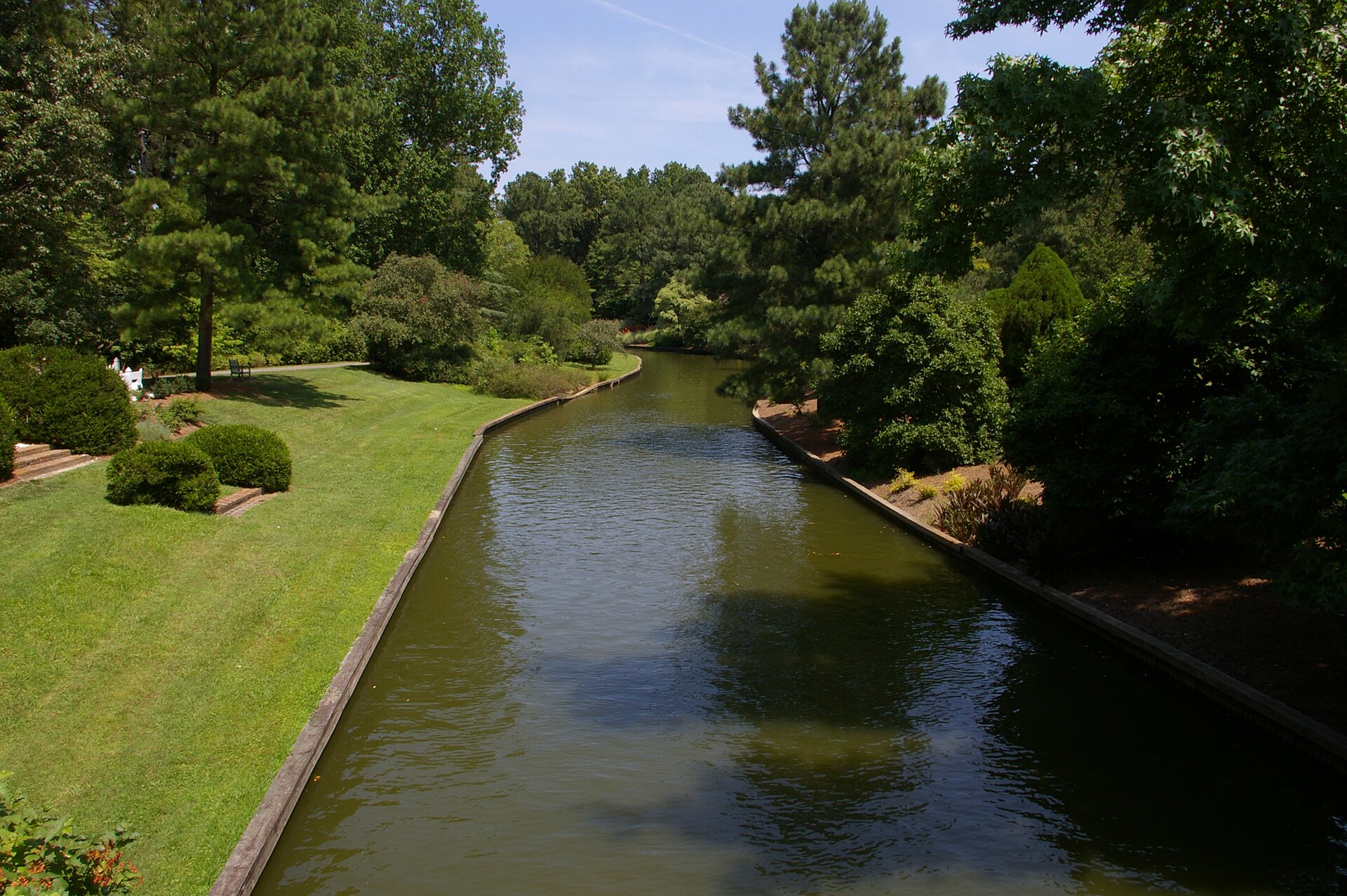 Manicured lawn, clipped shrubs and tall pines along a canal at Norfolk Botanical Garden